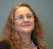 Woman with glasses smiles, wearing a black blazer and necklace.