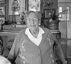An elderly woman with glasses sits indoors in front of a piano; she's wearing a sweater.