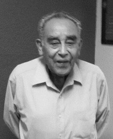 Elderly man, black and white photo. He is wearing a light button-down shirt. He has short hair, is smiling, and is indoors.