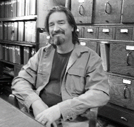 Man with beard smiling in a library, seated with arms crossed, books and filing cabinets in background.