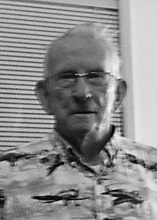 Elderly man wearing glasses and a patterned shirt, indoors.