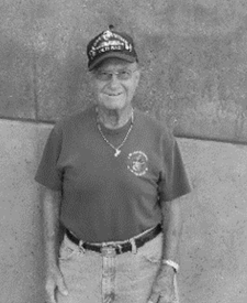 Elderly man in a cap and t-shirt, smiling, stands against a wall.