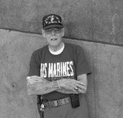 Elderly man in US Marines shirt and cap leans against wall, arms crossed, smoking.