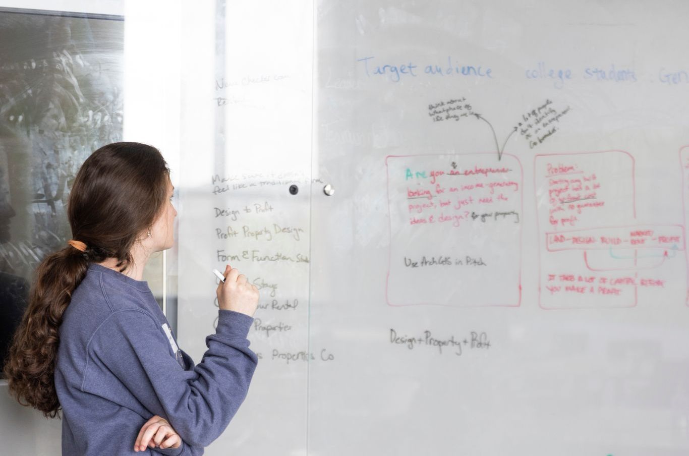 A person with dark hair in a ponytail stands in front of a whiteboard, writing and looking at the notes.