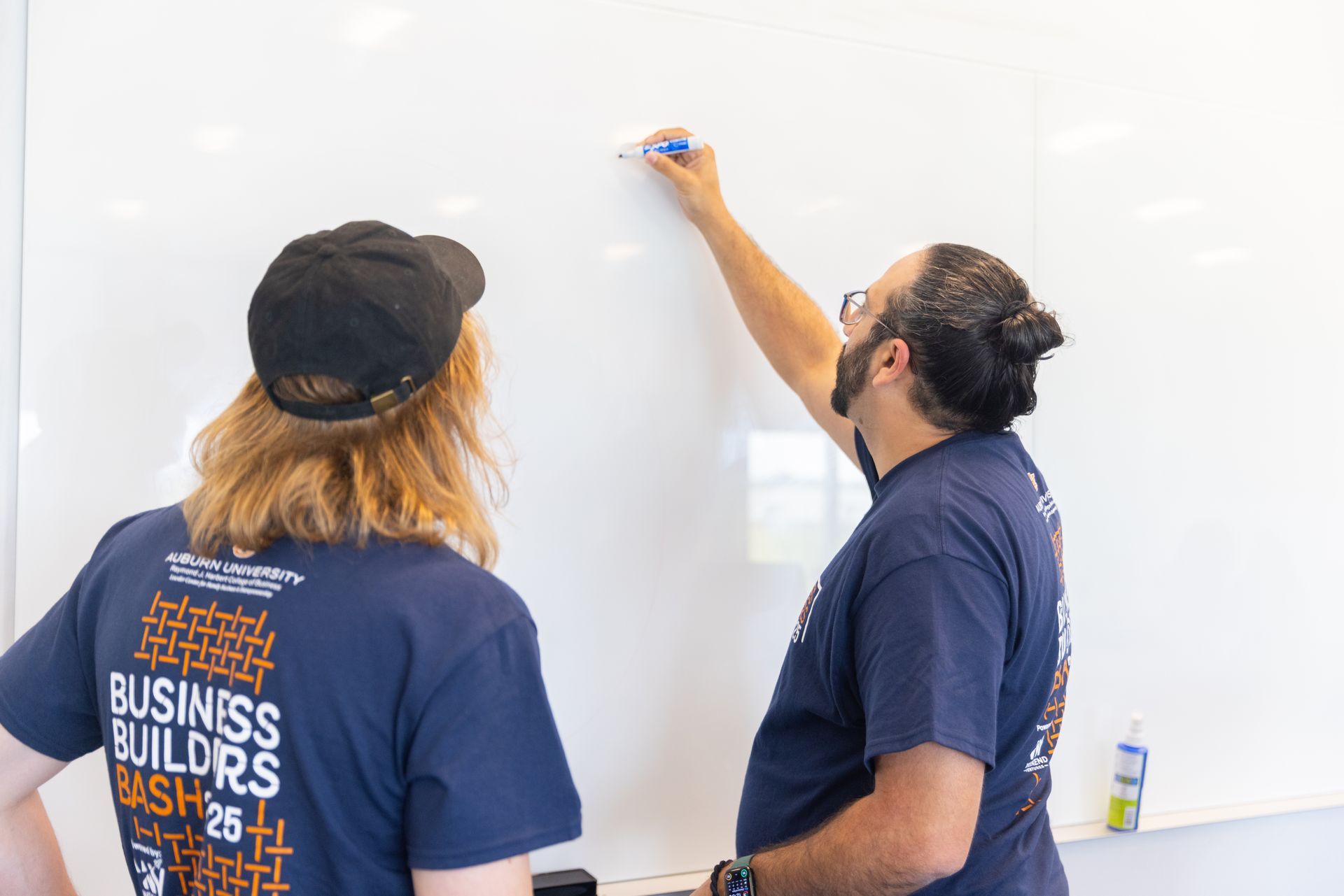 Two people in matching blue shirts stand in front of a whiteboard. One writes with a marker, the other watches. They appear to be indoors.