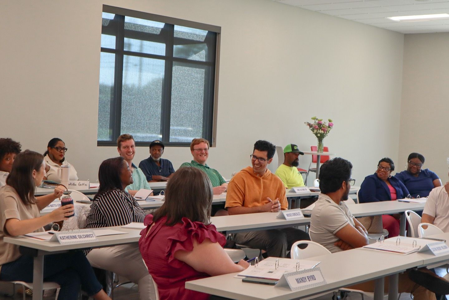 A group of diverse people seated at tables in a classroom setting. Some are smiling, others listening. A window is visible in the background.