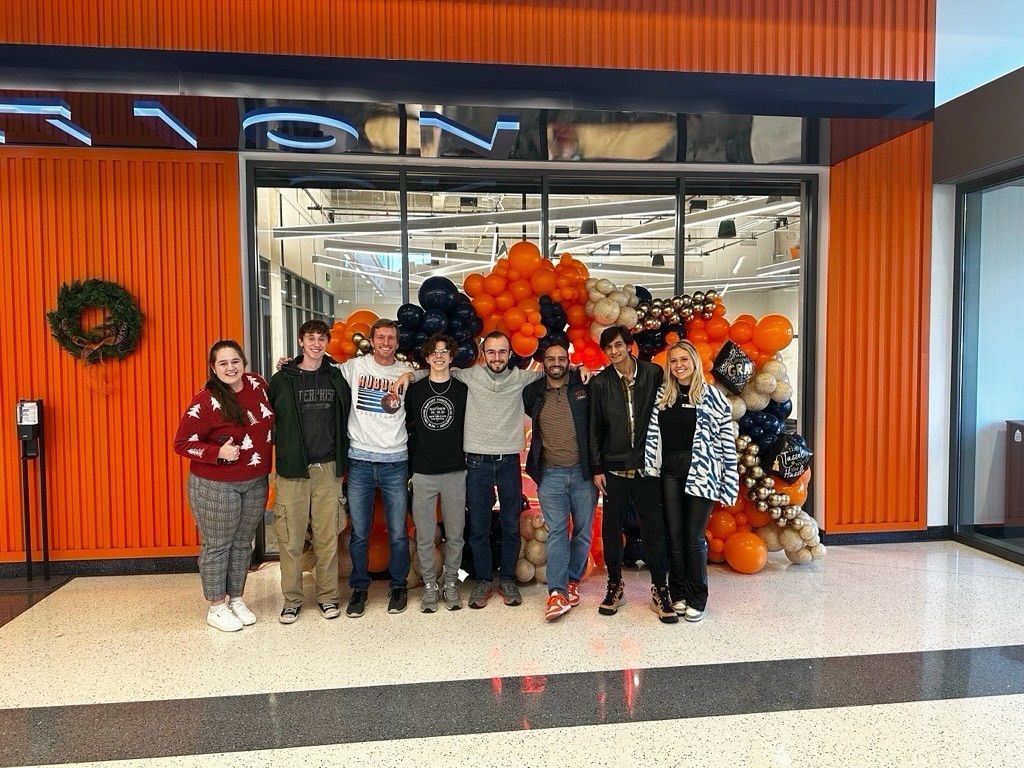Group of people standing in front of a store entrance with orange and black balloons, celebrating.