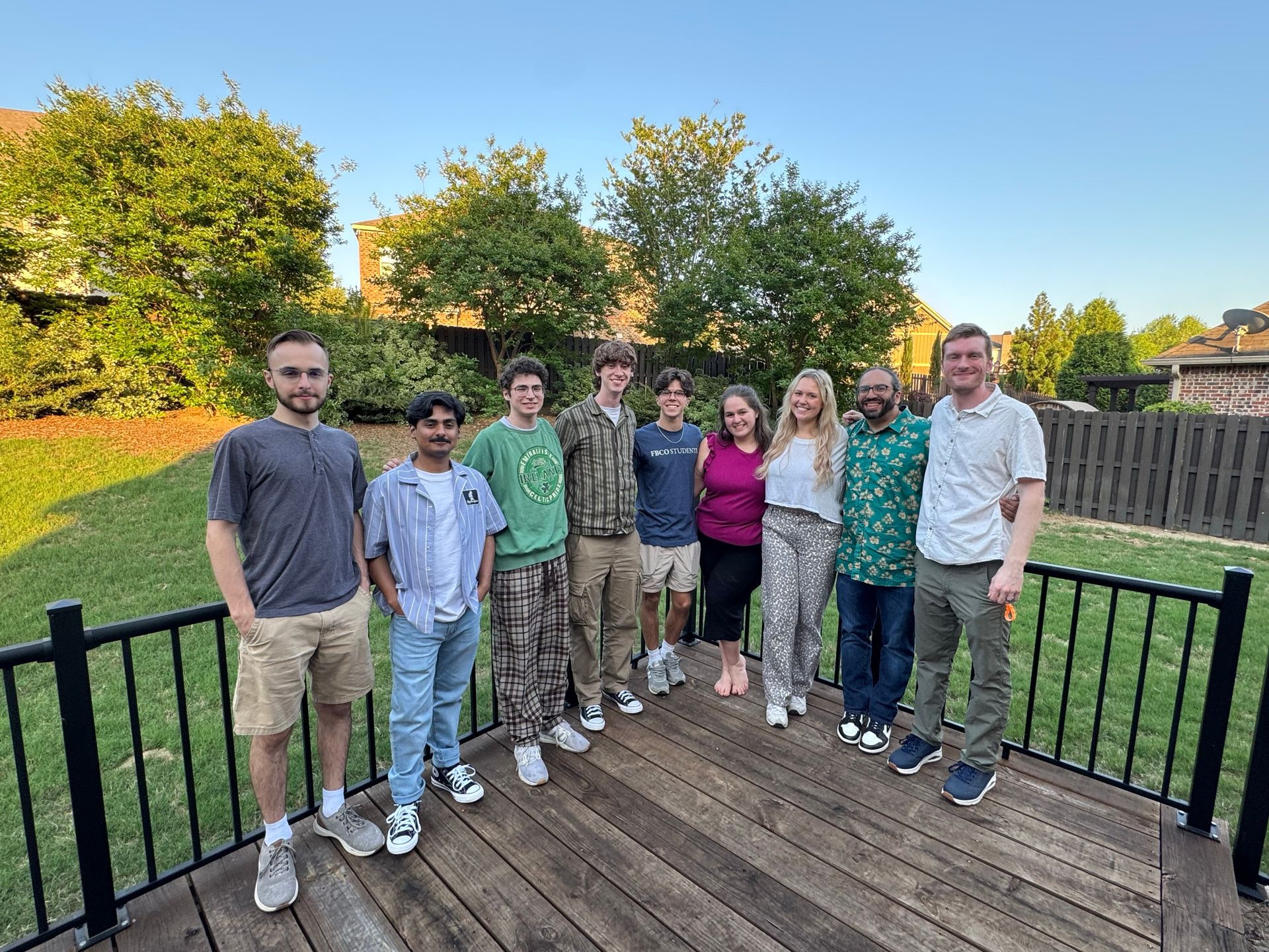 Group of ten people smiling on a wooden deck outside. They are standing in front of a fence and some trees, under a blue sky.
