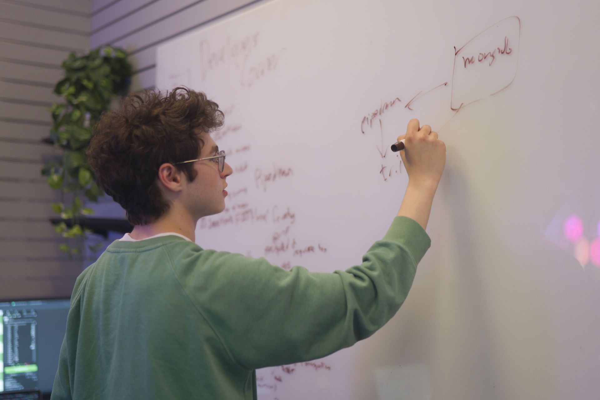 A young man with curly hair wearing glasses and a green sweater writes on a whiteboard, diagrams visible, in an office.