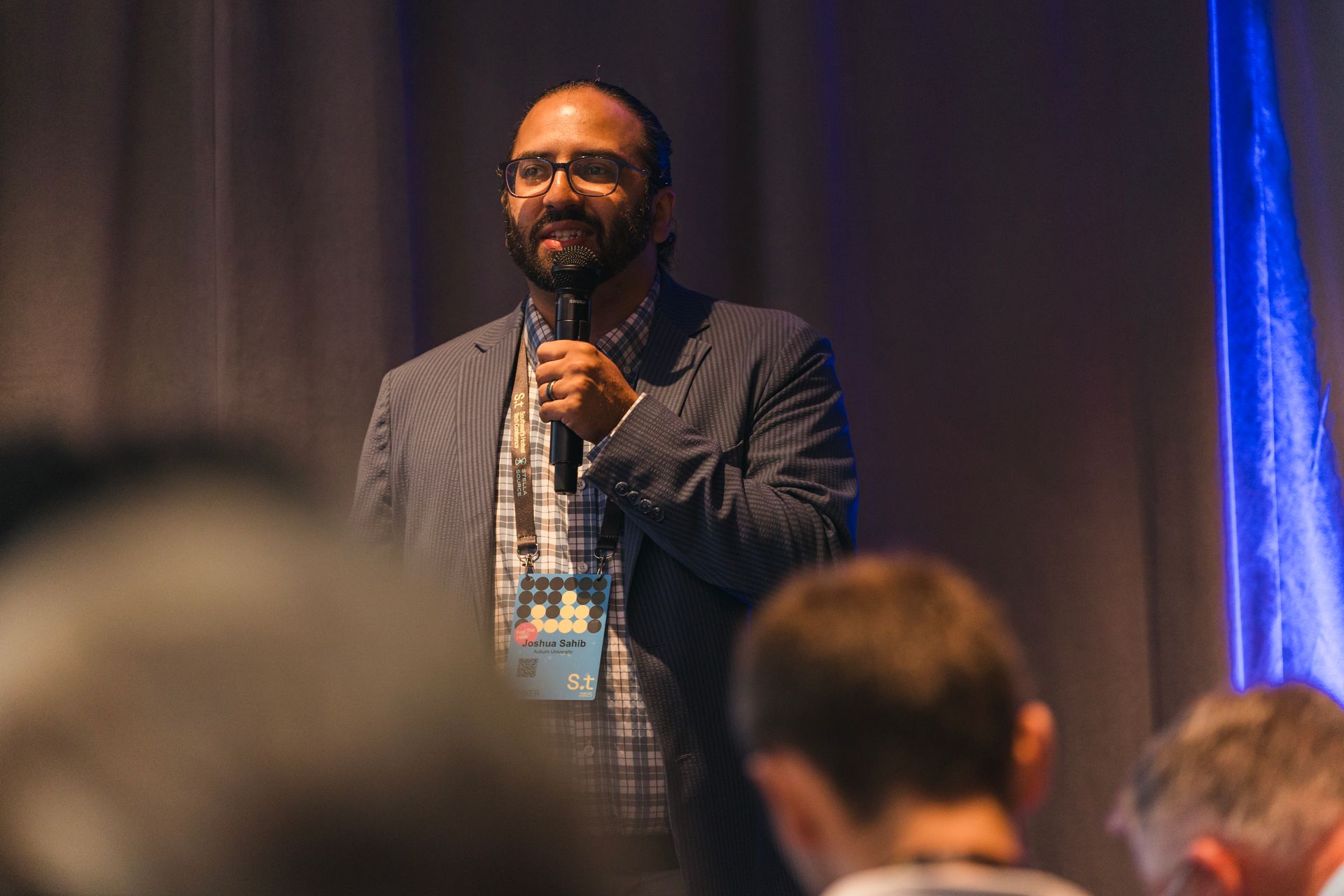 Man with beard and glasses speaks into a microphone on a stage, wearing a blazer over a plaid shirt.