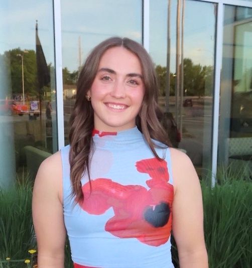 Smiling woman in a light blue sleeveless top with pink accents, standing outside a glass building.