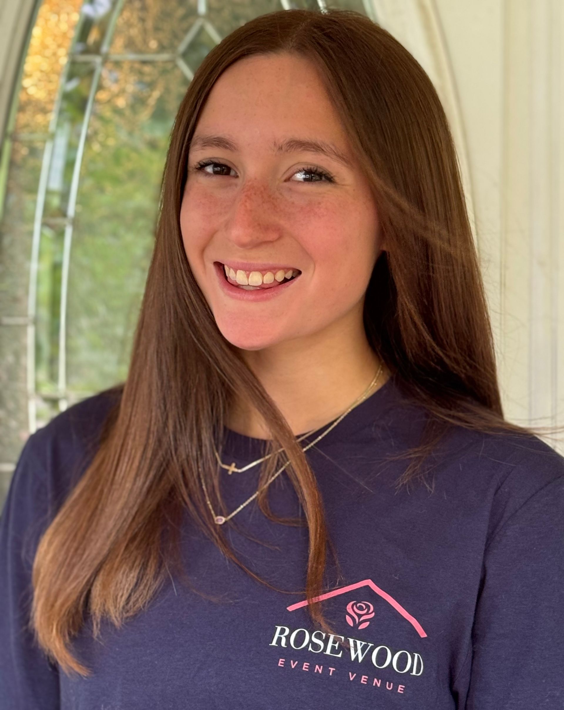 Smiling young person in a navy Rosewood shirt indoors by a window.
