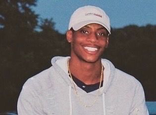 Smiling person in a light gray hoodie and white cap outdoors at dusk.