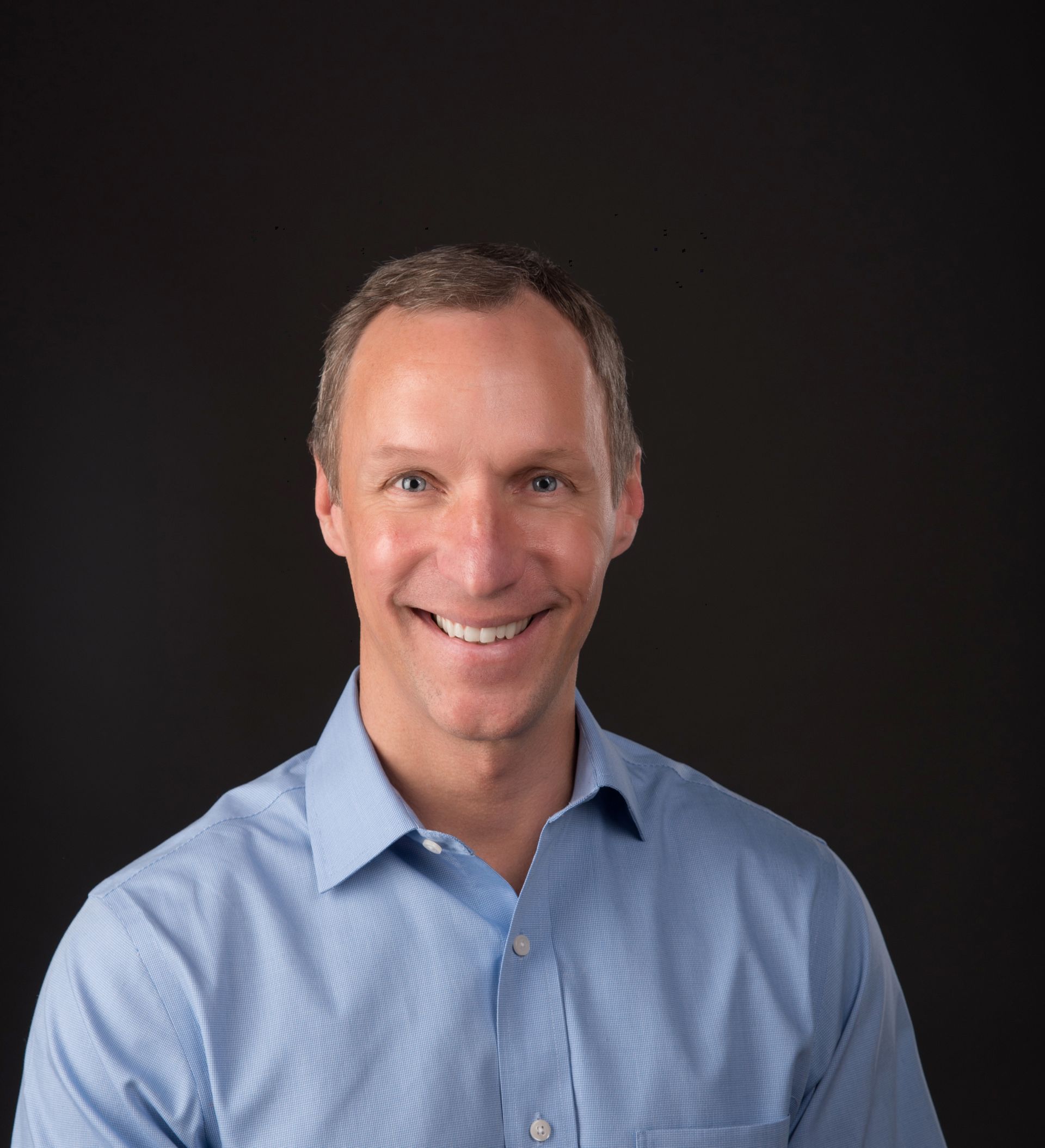Man smiling in a blue button-down shirt against a dark background.