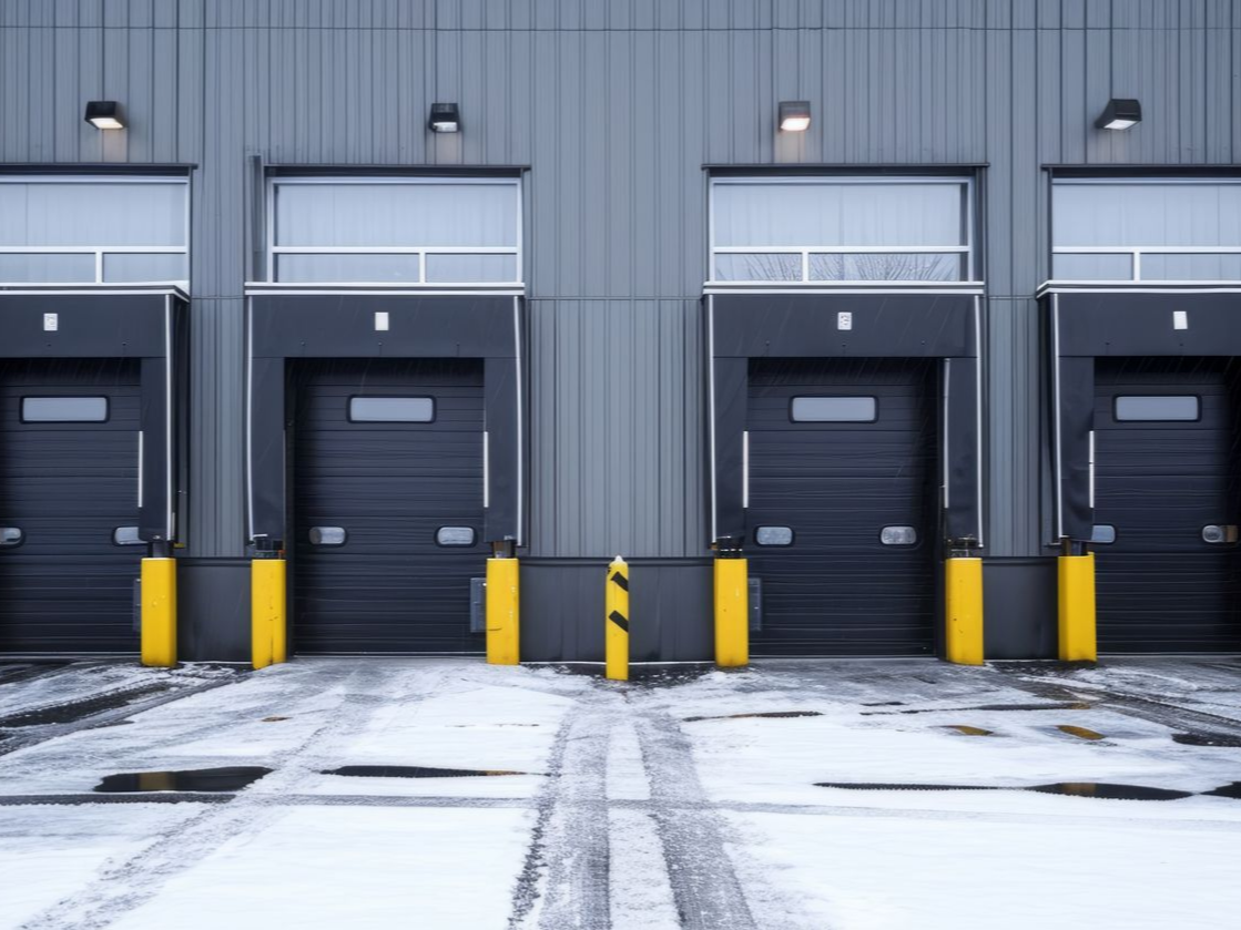 Loading docks in a row, snow-covered ground, black doors with yellow bollards.