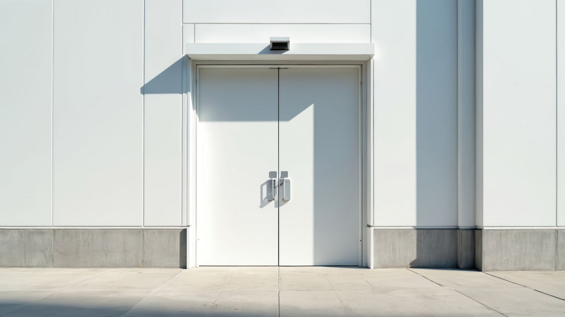 White double doors on a white building, casting shadows onto a concrete sidewalk.