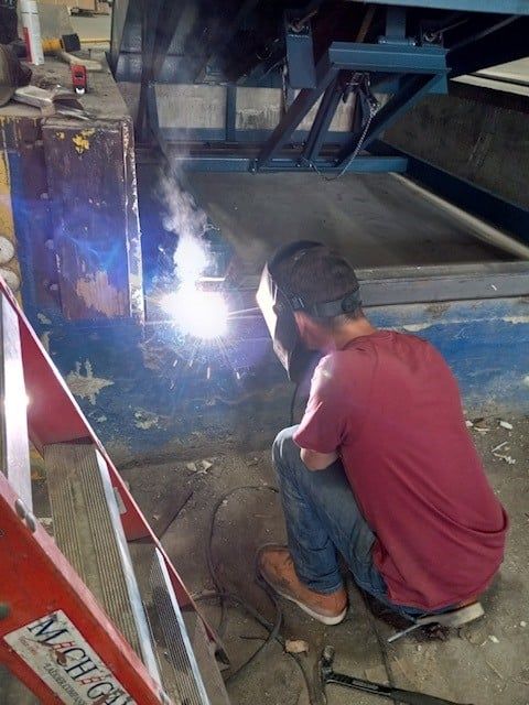 Person welding metal, wearing a mask, squatting in a workshop. Bright blue light and smoke.