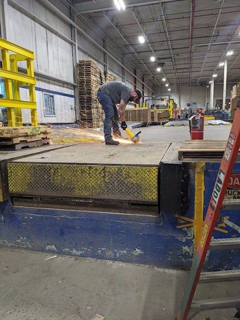 Person using a grinder on a loading dock, sparks flying. Warehouse setting, pallets and ladder present.