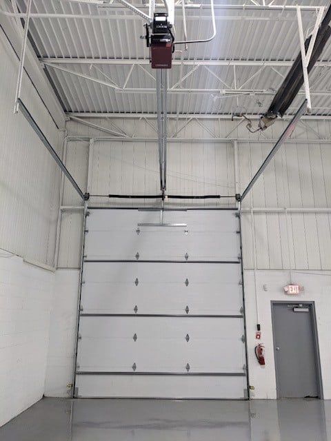 Large white commercial garage door, closed, with overhead opener in a warehouse setting.