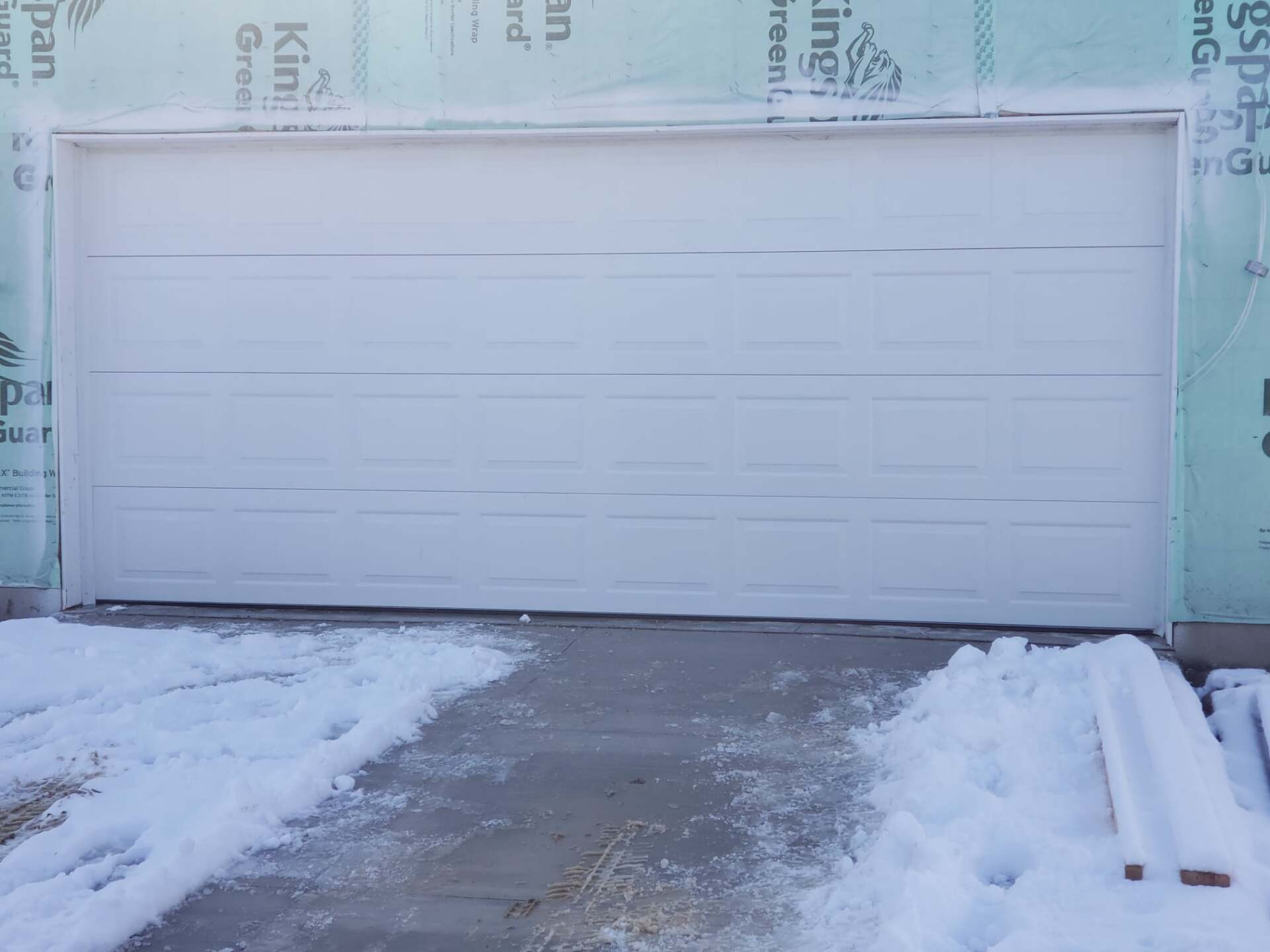 White garage door, partially closed, in snowy setting; construction materials nearby.