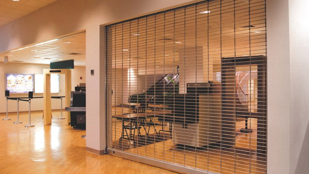 A clear, retractable security gate separates a room from a hallway. The room has a piano and tables.