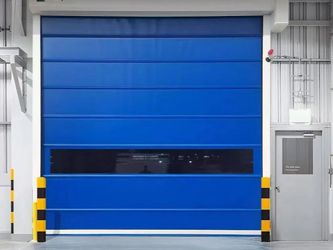 Blue industrial roll-up door with a black window. Building interior with a side door and yellow and black bollards.