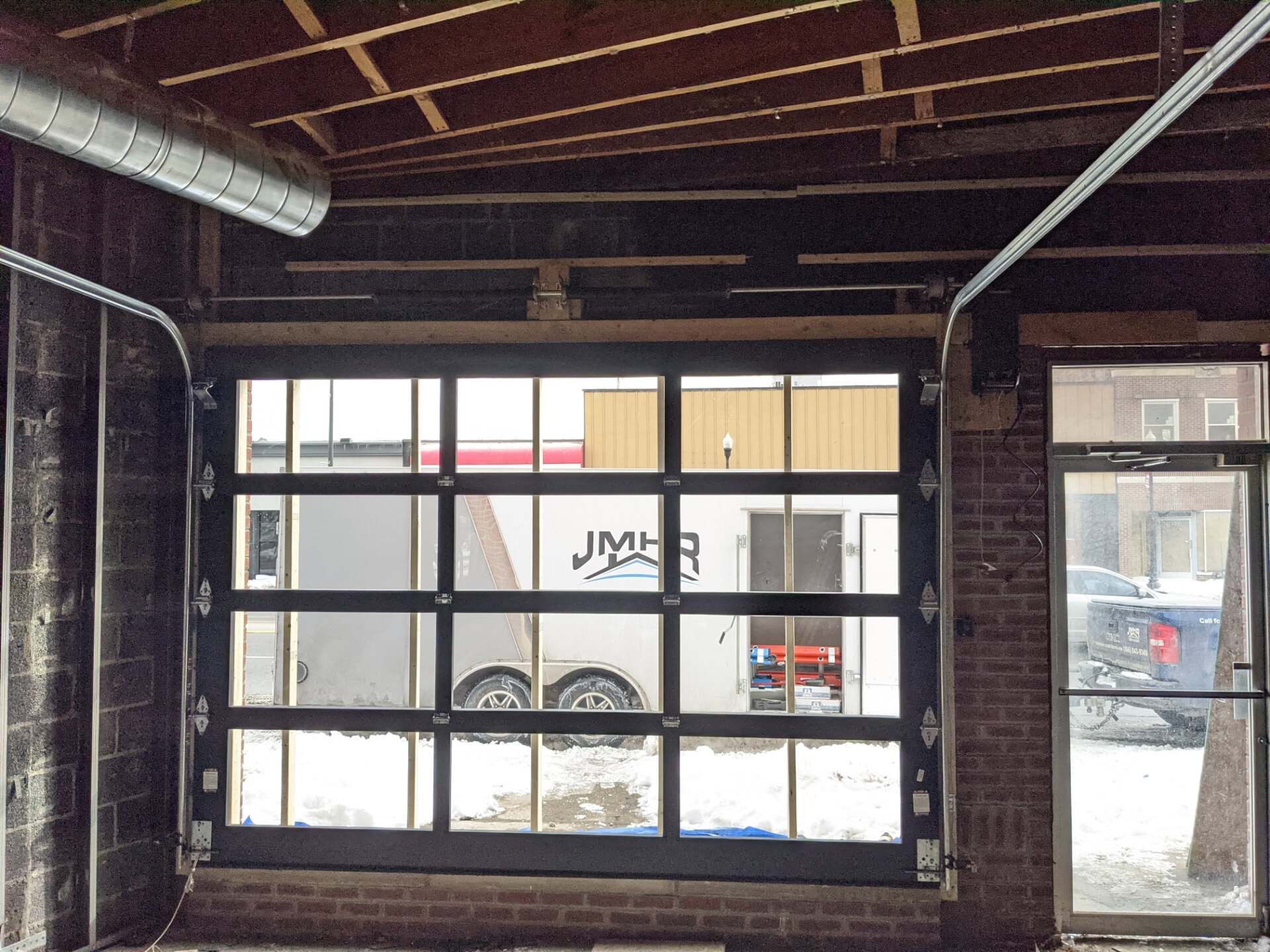 Glass garage door, partially open, revealing a trailer and a window. Building interior with exposed brick and ductwork.