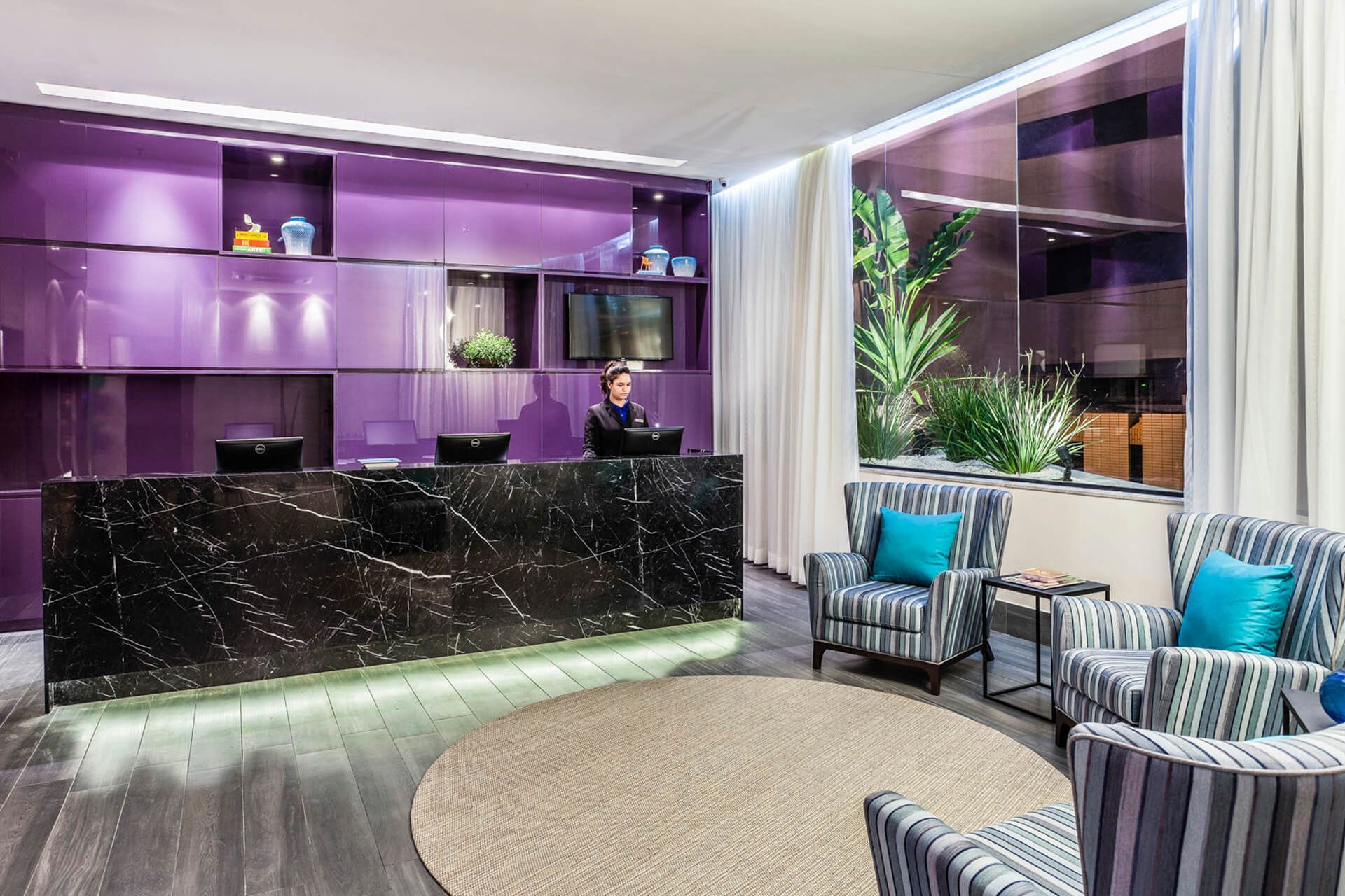 Um lobby de hotel com paredes roxas, um balcão de recepção, cadeiras e um tapete redondo.