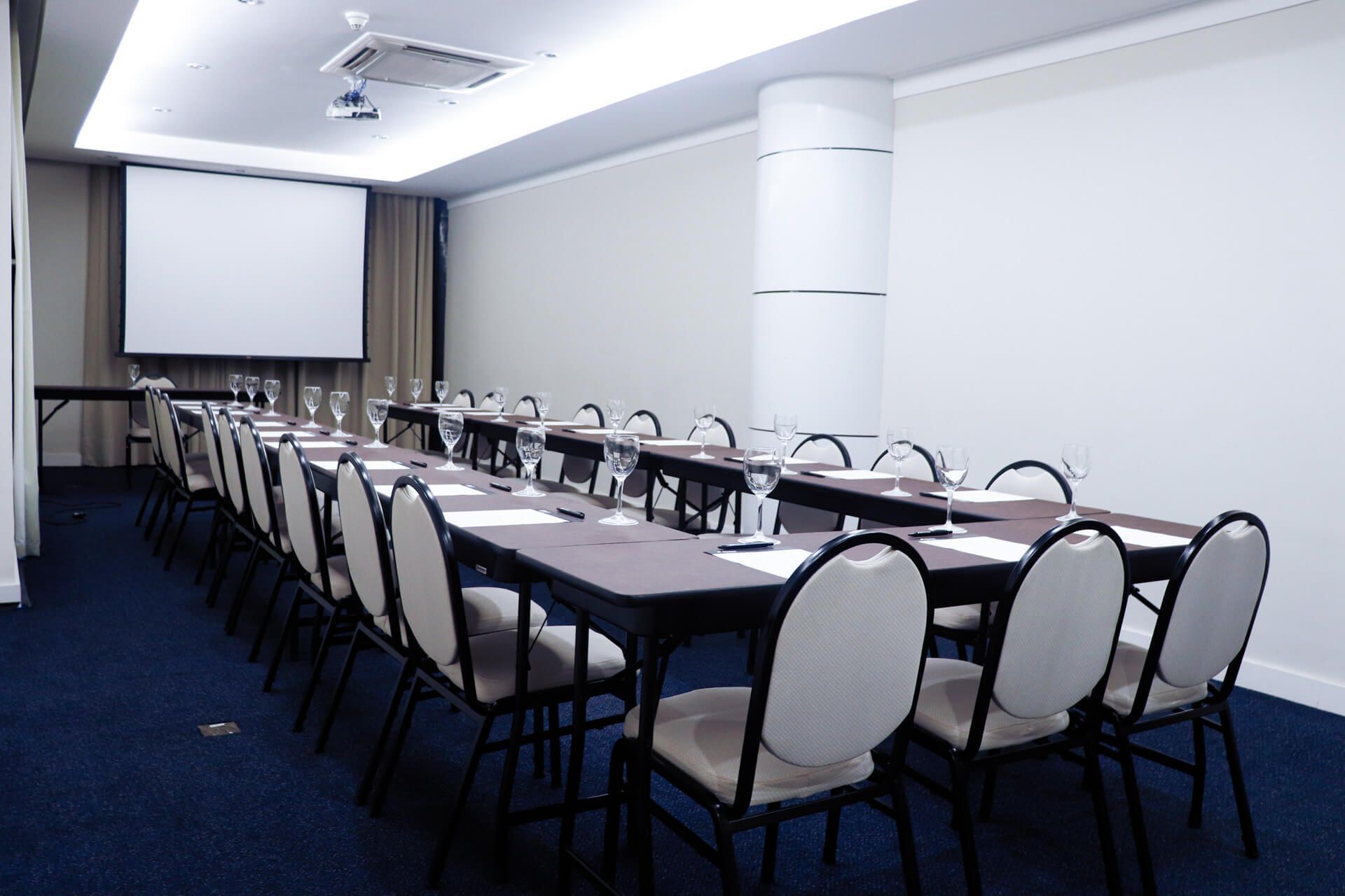 Uma sala de conferências com mesas e cadeiras e uma tela de projetor.