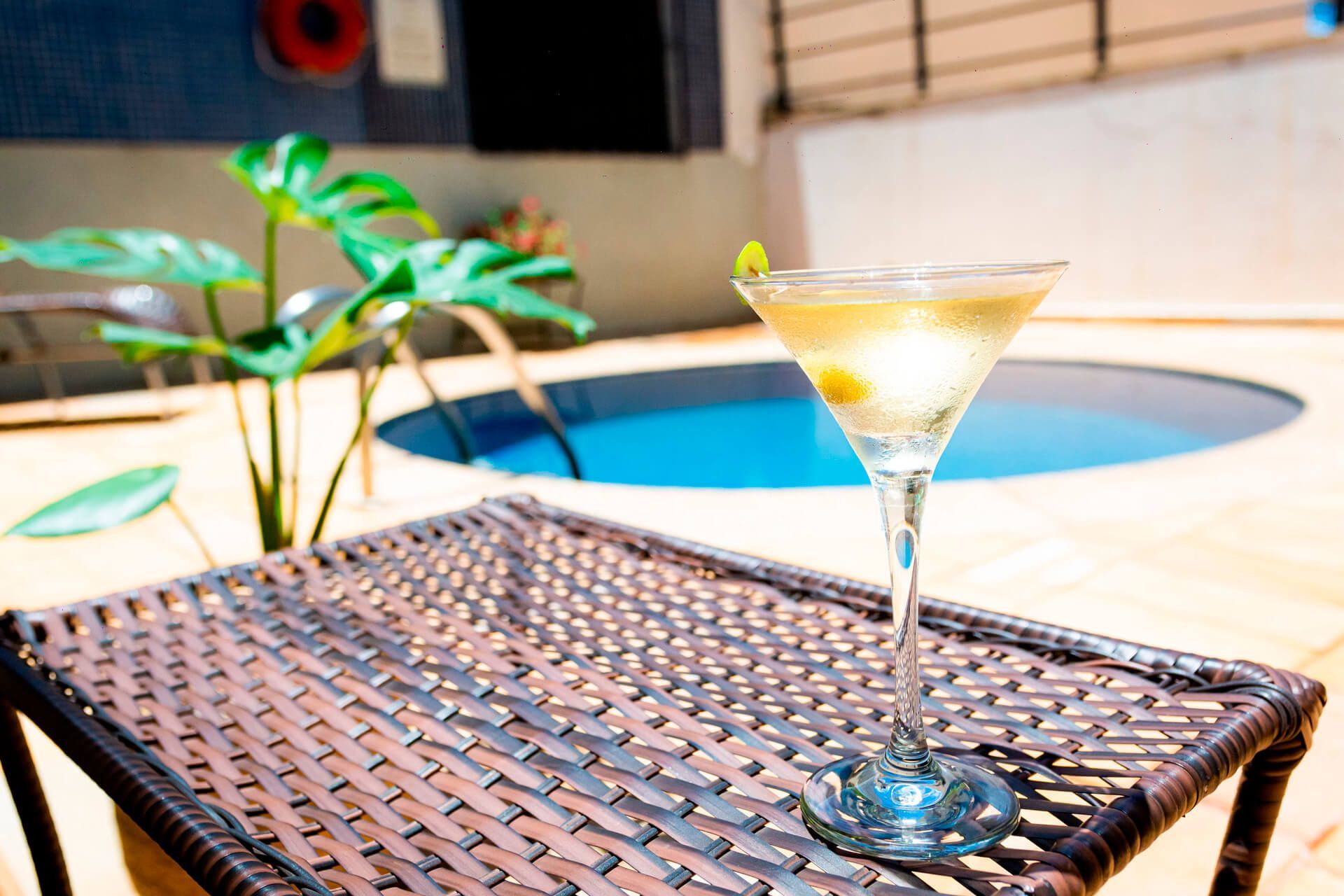 Uma taça de martini está sobre uma mesa de vime em frente a uma piscina.