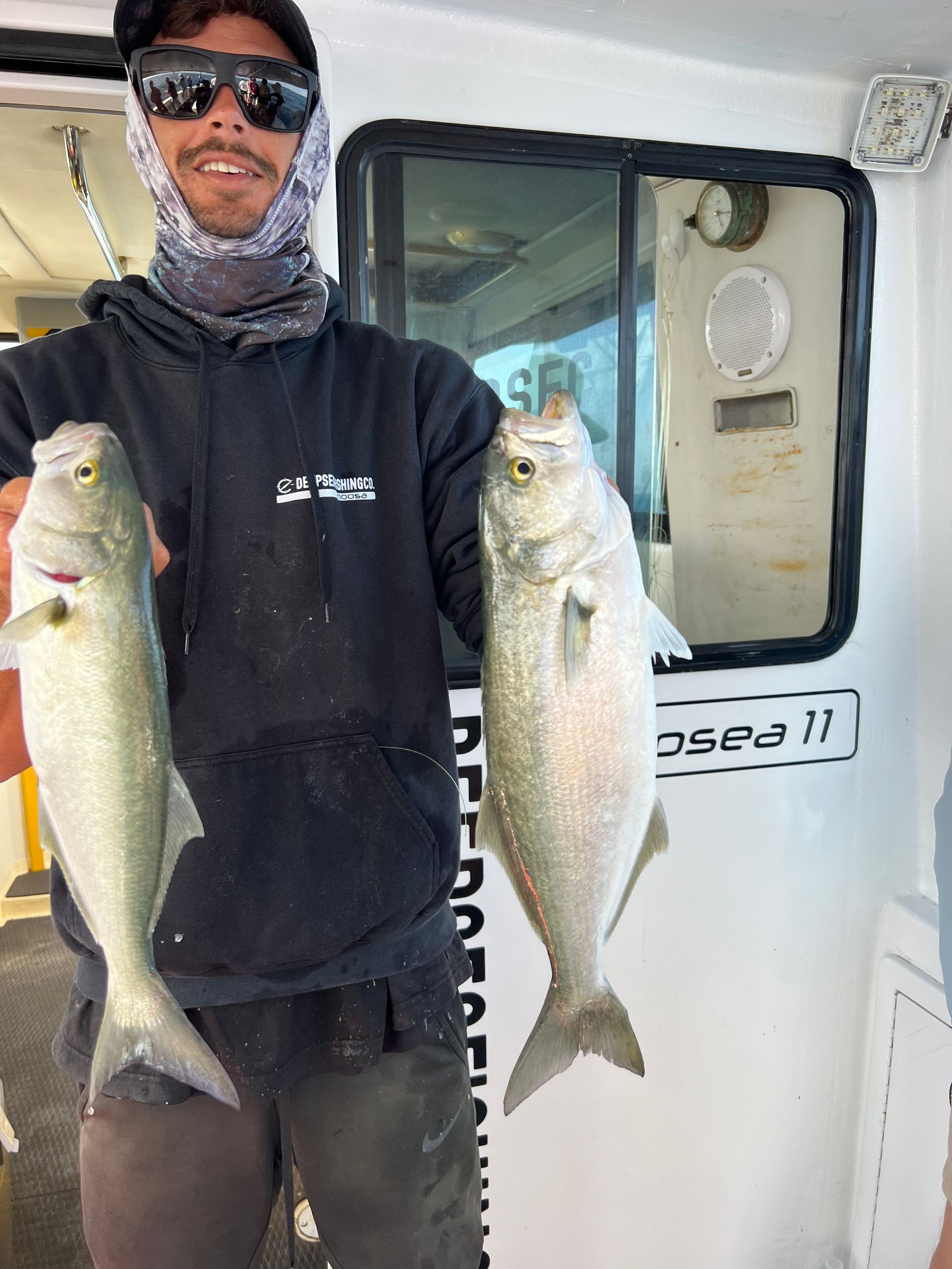 A man wearing sunglasses is holding two fish in front of a boat.