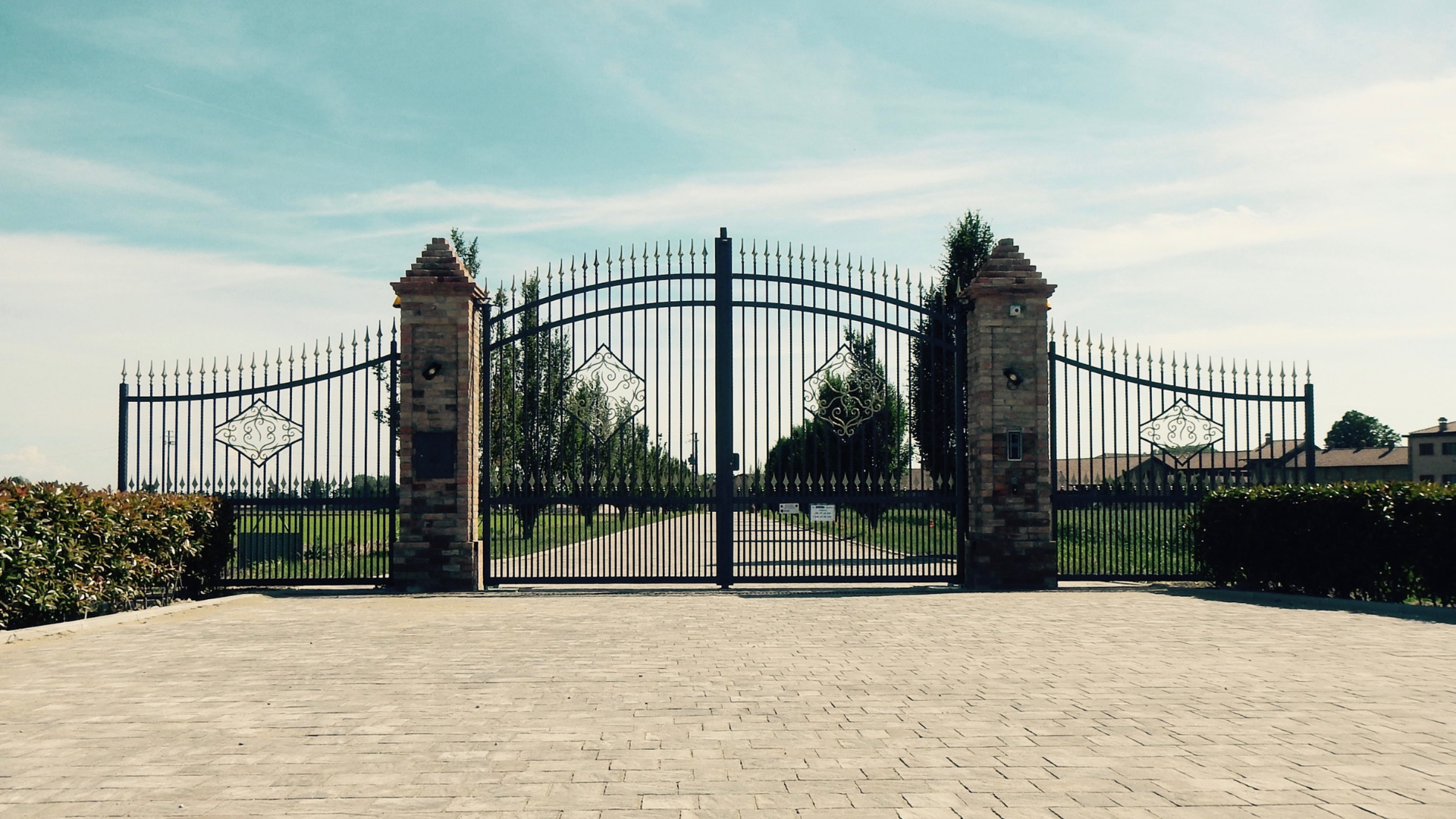 Ornate black iron gates open to a long driveway, leading to a property under a bright blue sky.