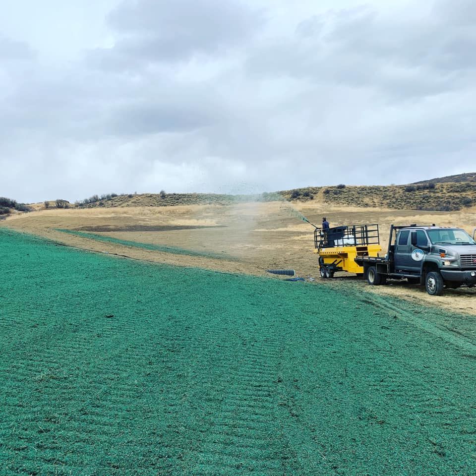 A truck towing a hydroseeder sprays a green slurry on a hillside, covering a field of green netting.