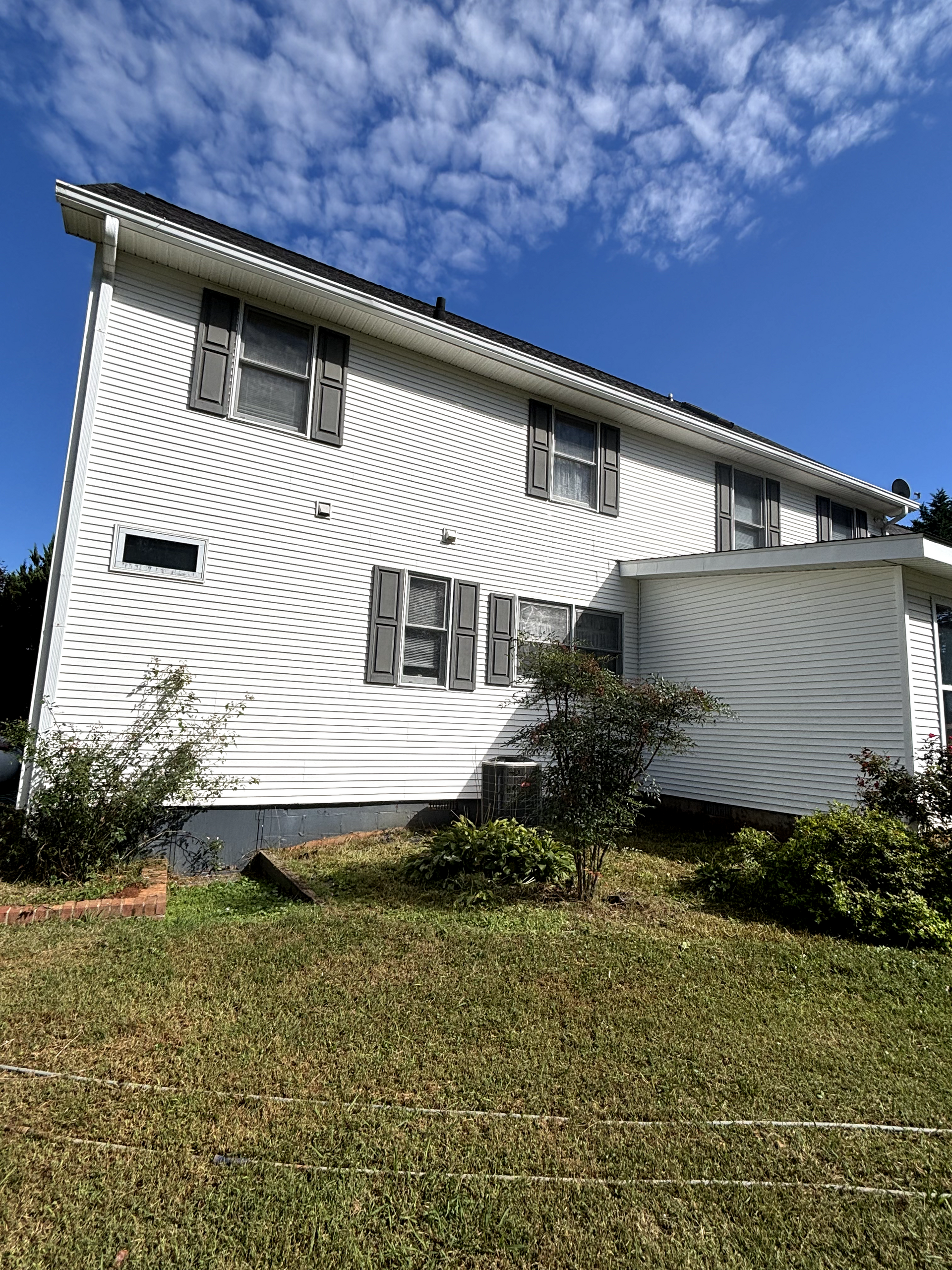 White two-story house with gray shutters against a blue sky, surrounded by green grass and bushes.
