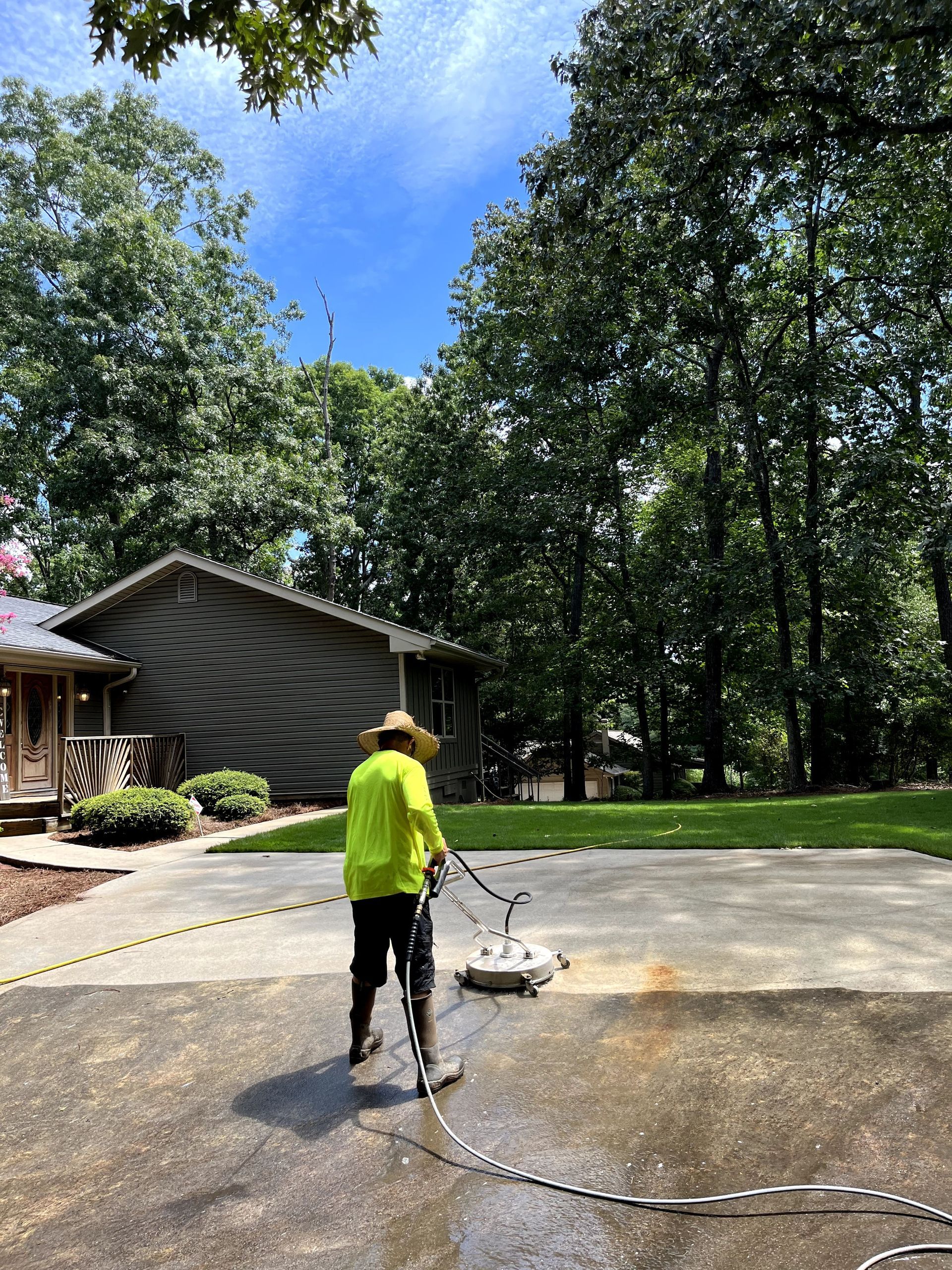Person in neon yellow shirt power washing a concrete driveway on a sunny day. Trees and a house in the background.
