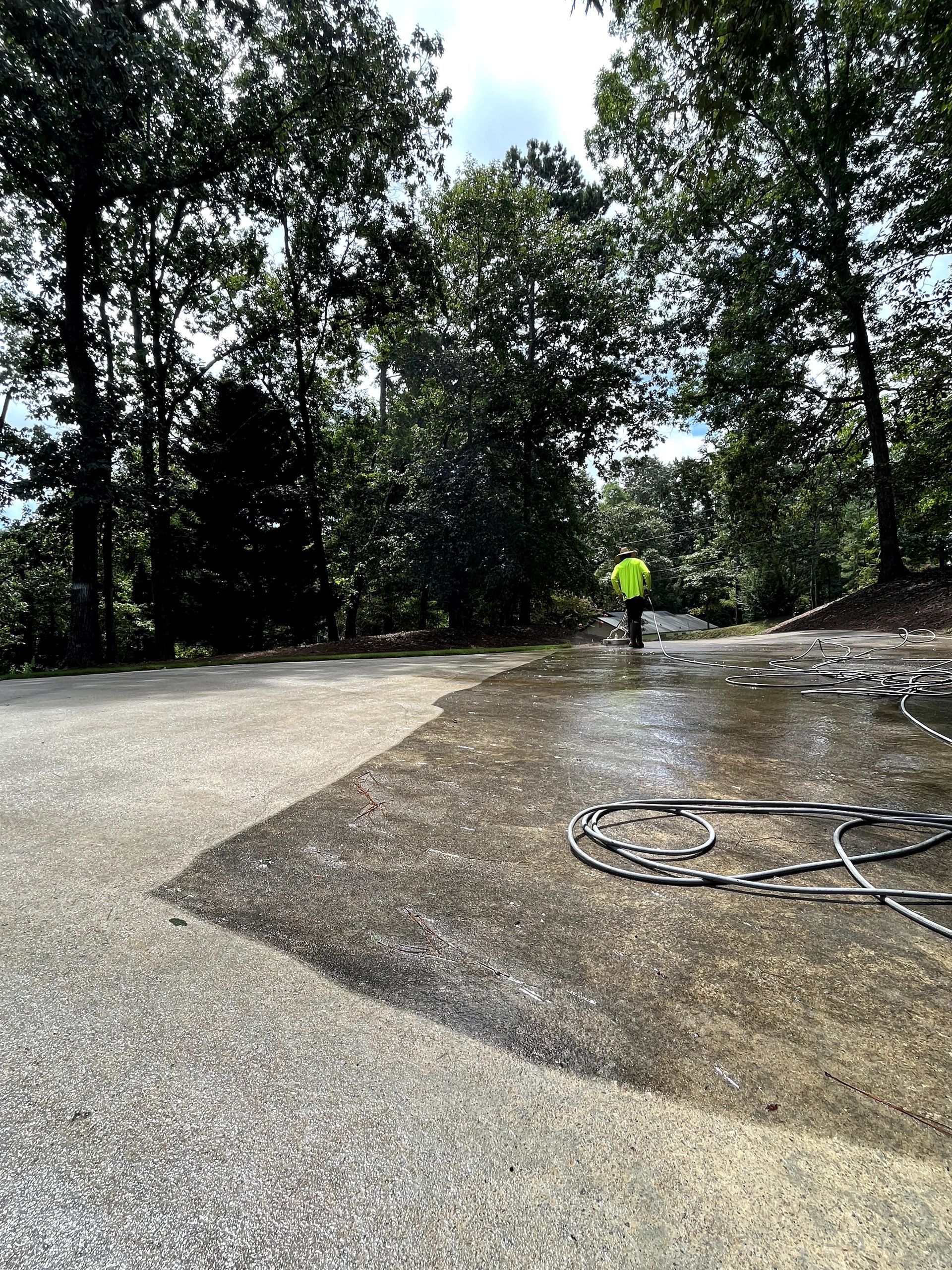 Person power washing a concrete driveway with trees in the background. Wet surface and hose visible.