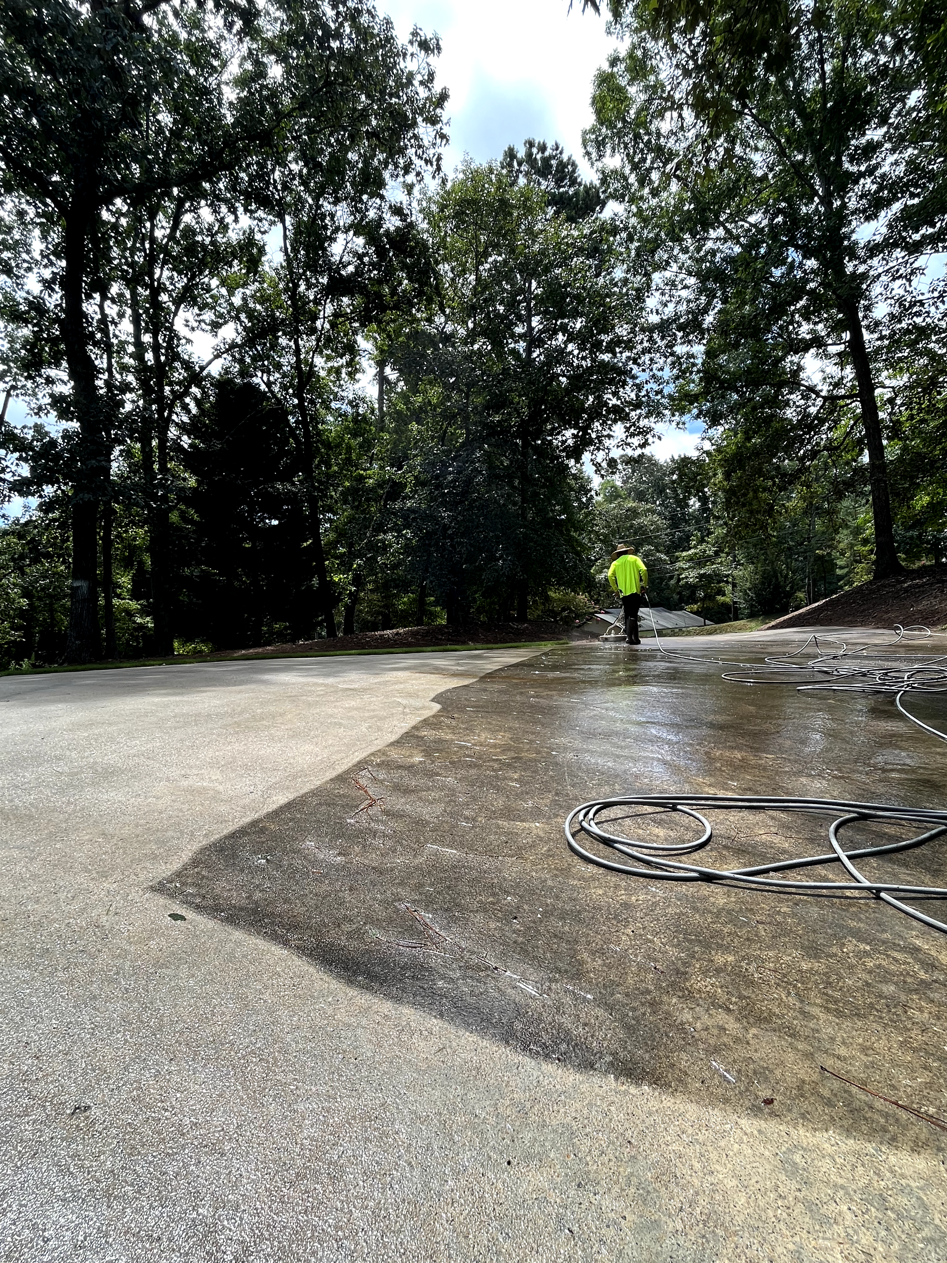 Person in yellow vest pressure washing a wet concrete driveway, surrounded by trees.
