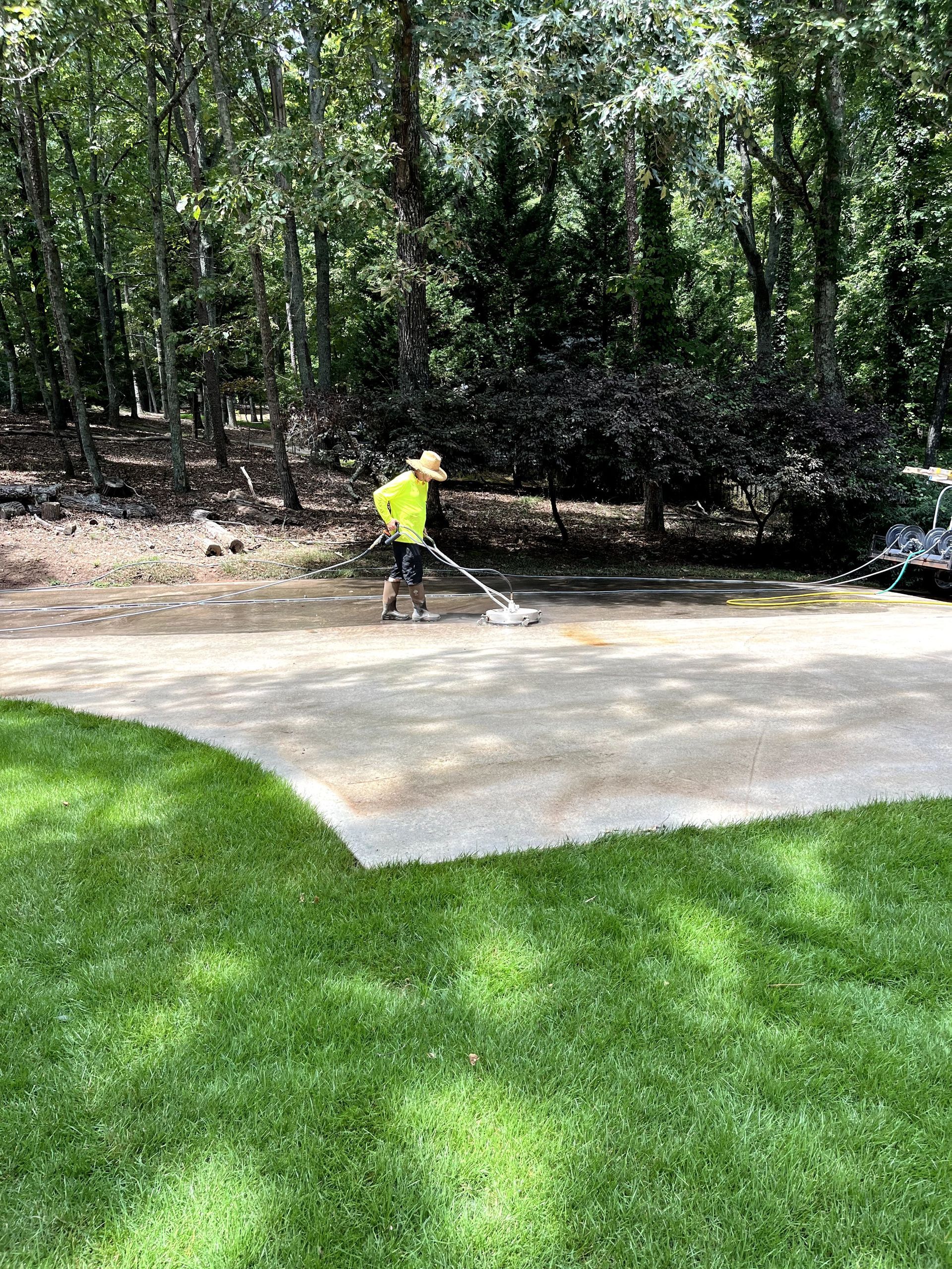 Person in yellow safety vest cleaning a concrete driveway with trees in the background. Green grass on the left.