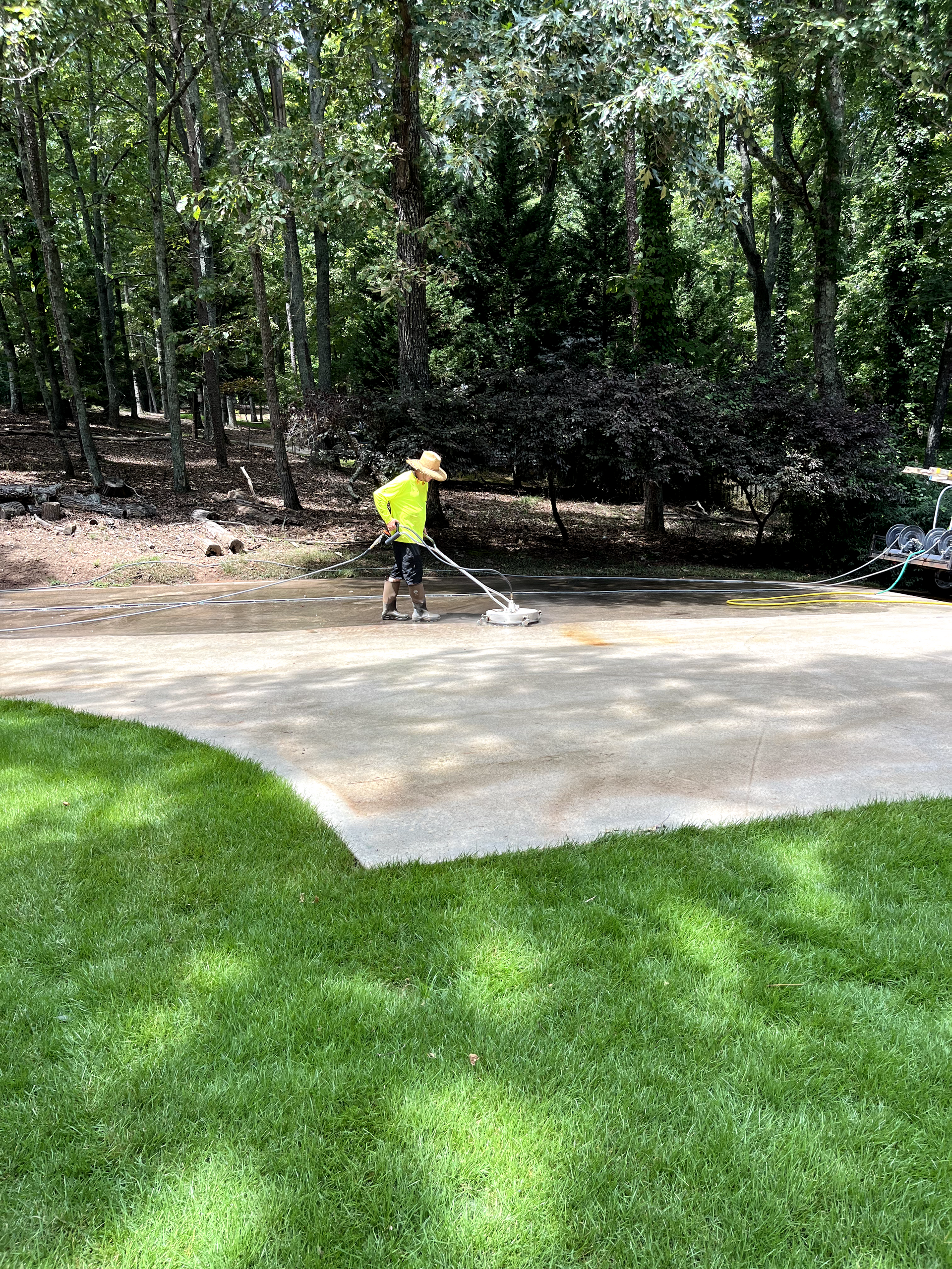 Person in yellow shirt power washing a concrete driveway near a grassy lawn and trees.