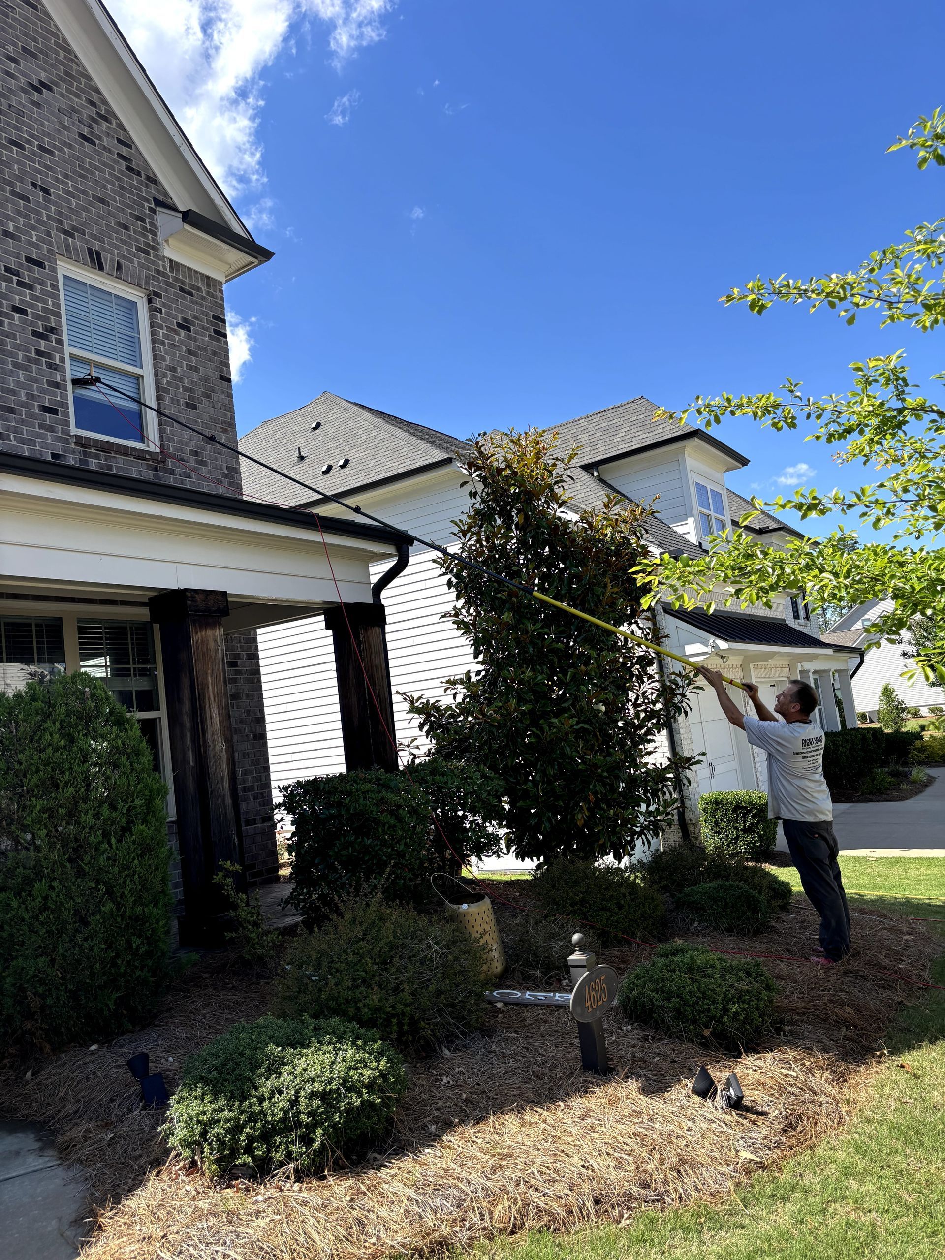 Man trimming branches near a house with brick and a dark roof under a blue sky.