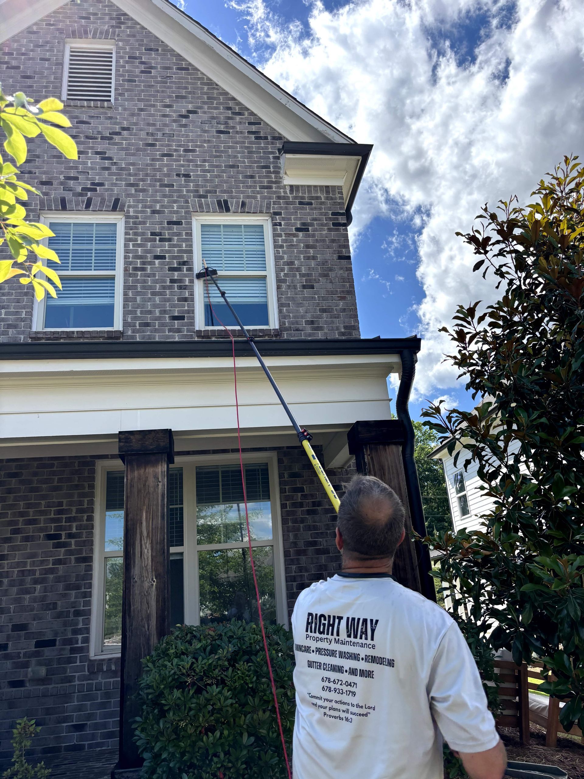 Man cleaning a house gutter with a long pole. A brick house, blue sky, and foliage are in the background.