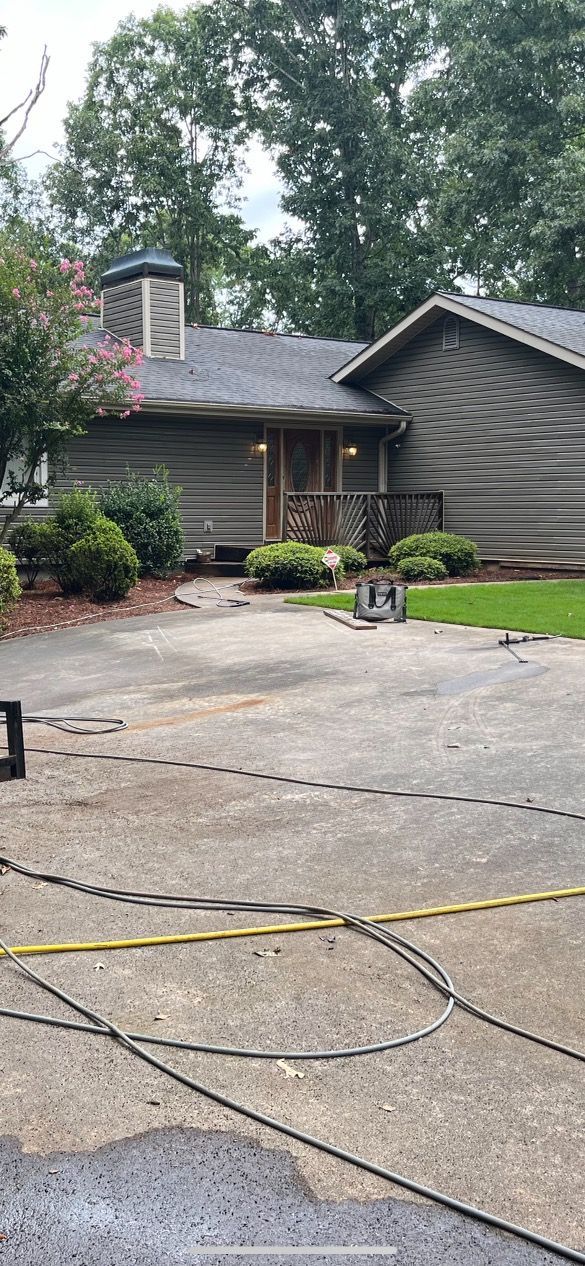 A single-story wood-sided house with a dark roof and a chimney sits behind a paved driveway and some landscaping.