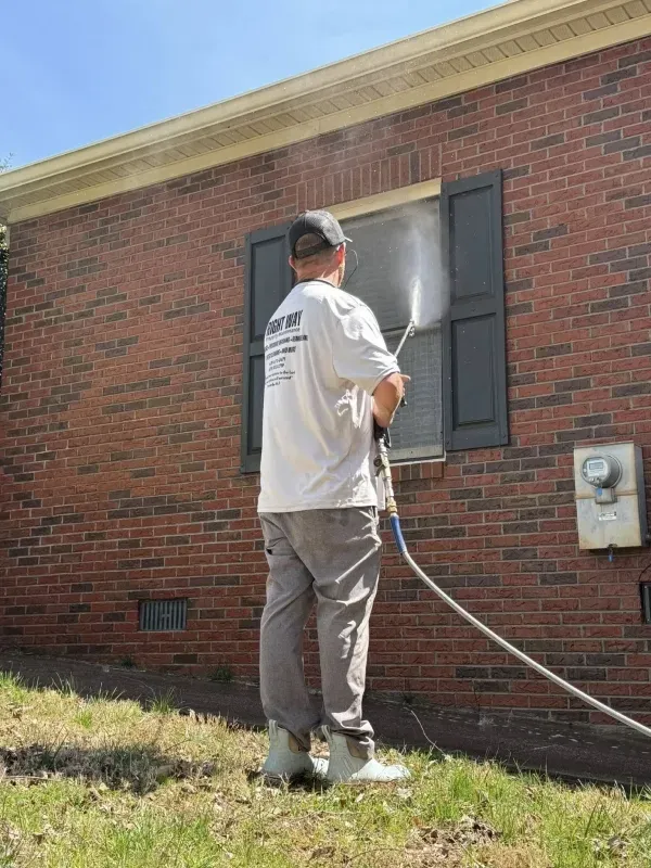 Man power washing a window on a brick building with black shutters.