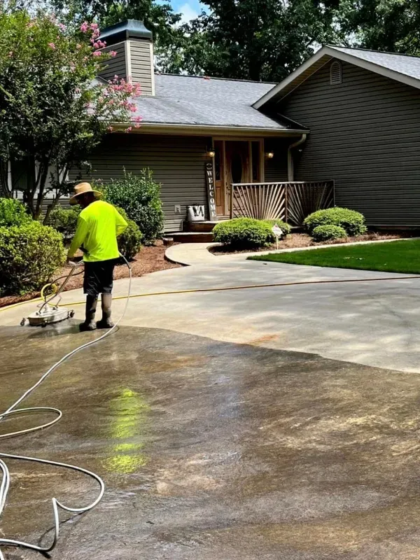 Person pressure washing a driveway next to a house with a porch and bushes.
