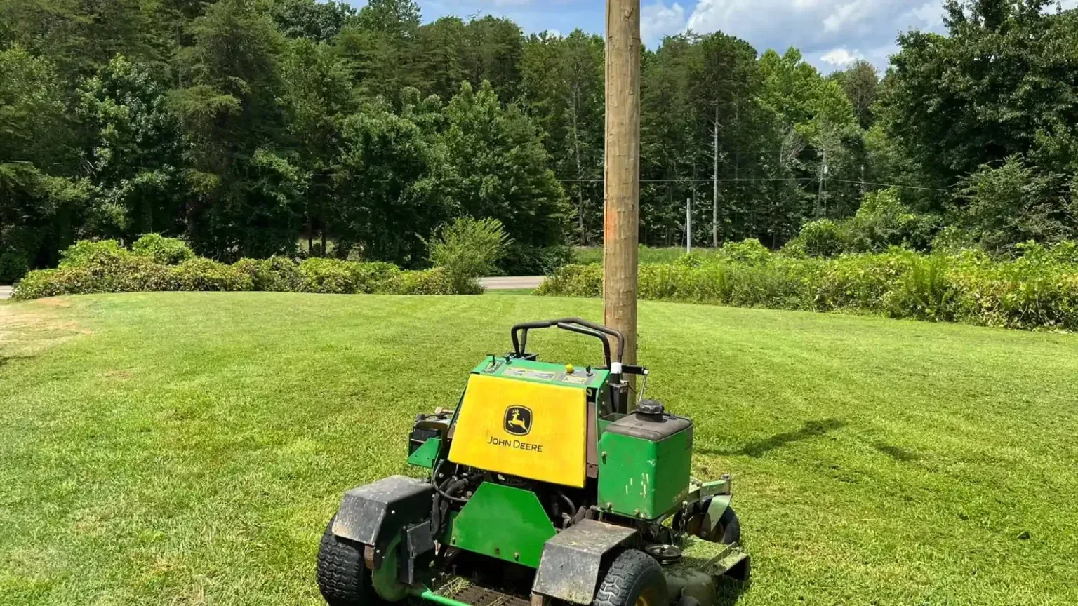 A John Deere utility vehicle with a tall pole stands on a green lawn with a tree line background.