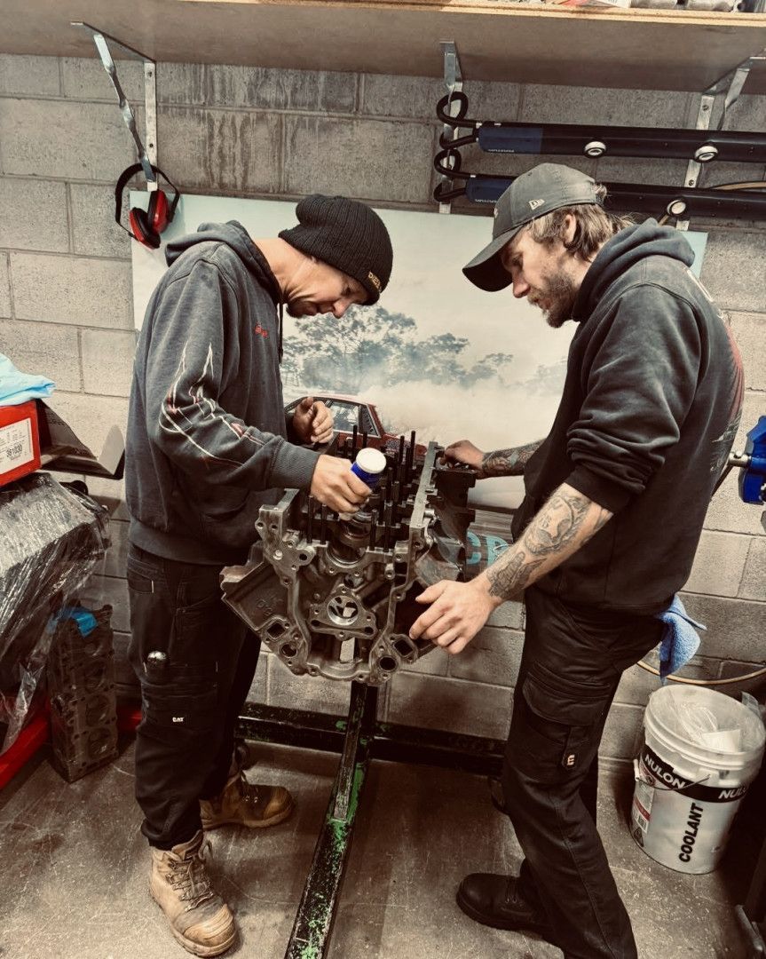 Two men in a garage working on an engine block. One sprays lubricant.