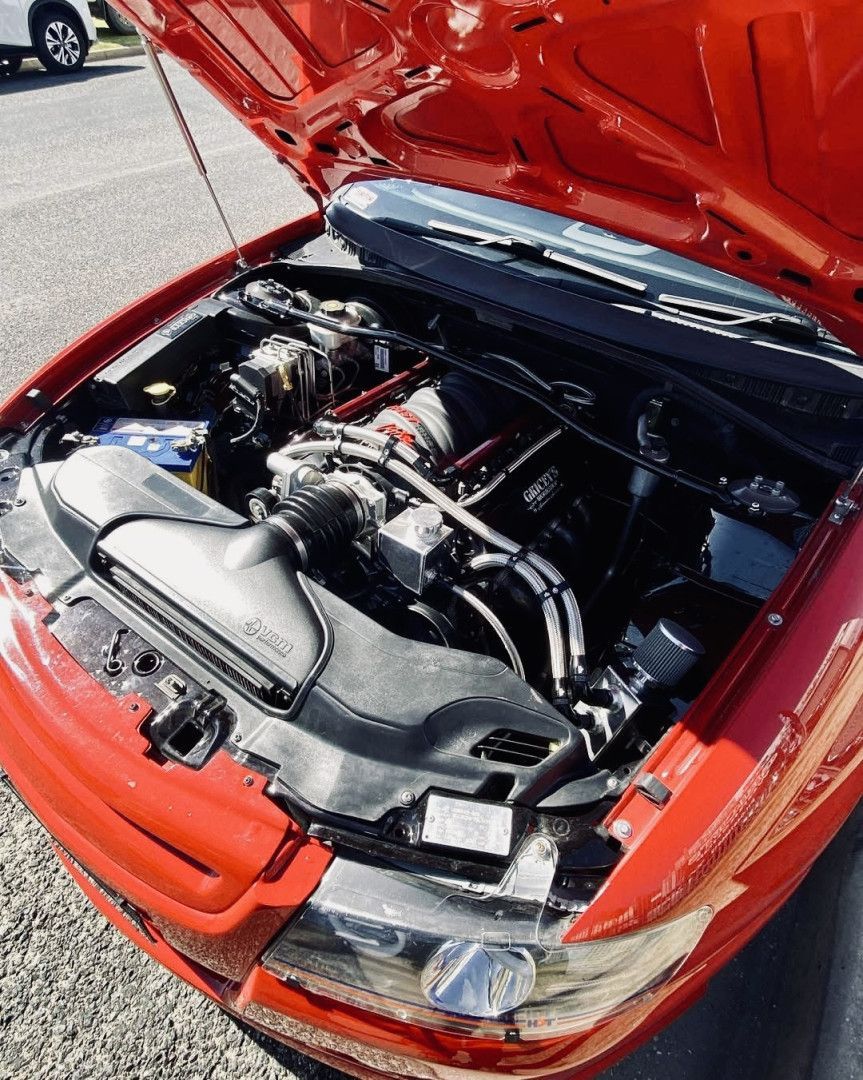 Red car engine bay, open hood. Black engine components, chrome trim, and blue battery.