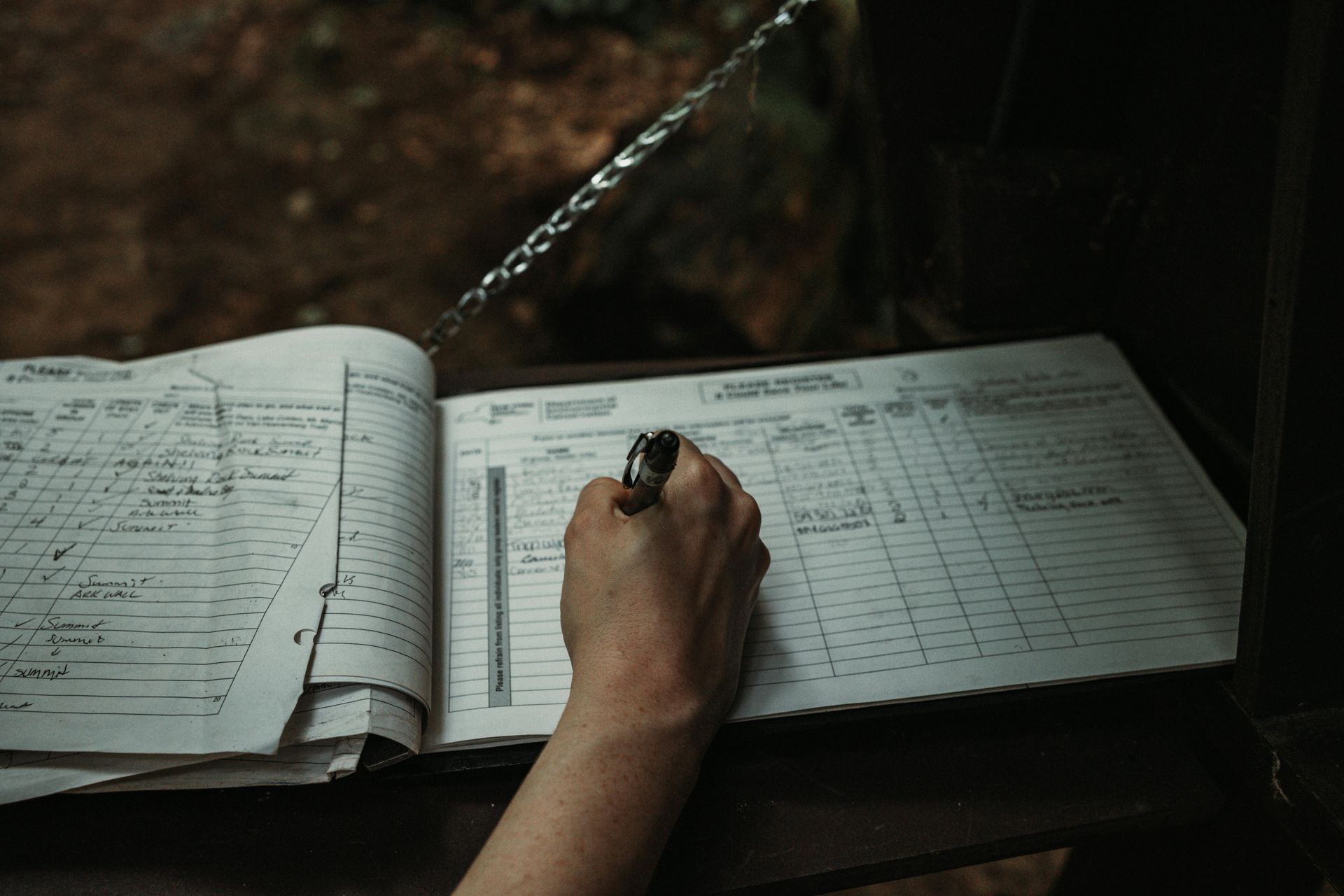 Person writing in a large ledger outdoors; silver water runs above.