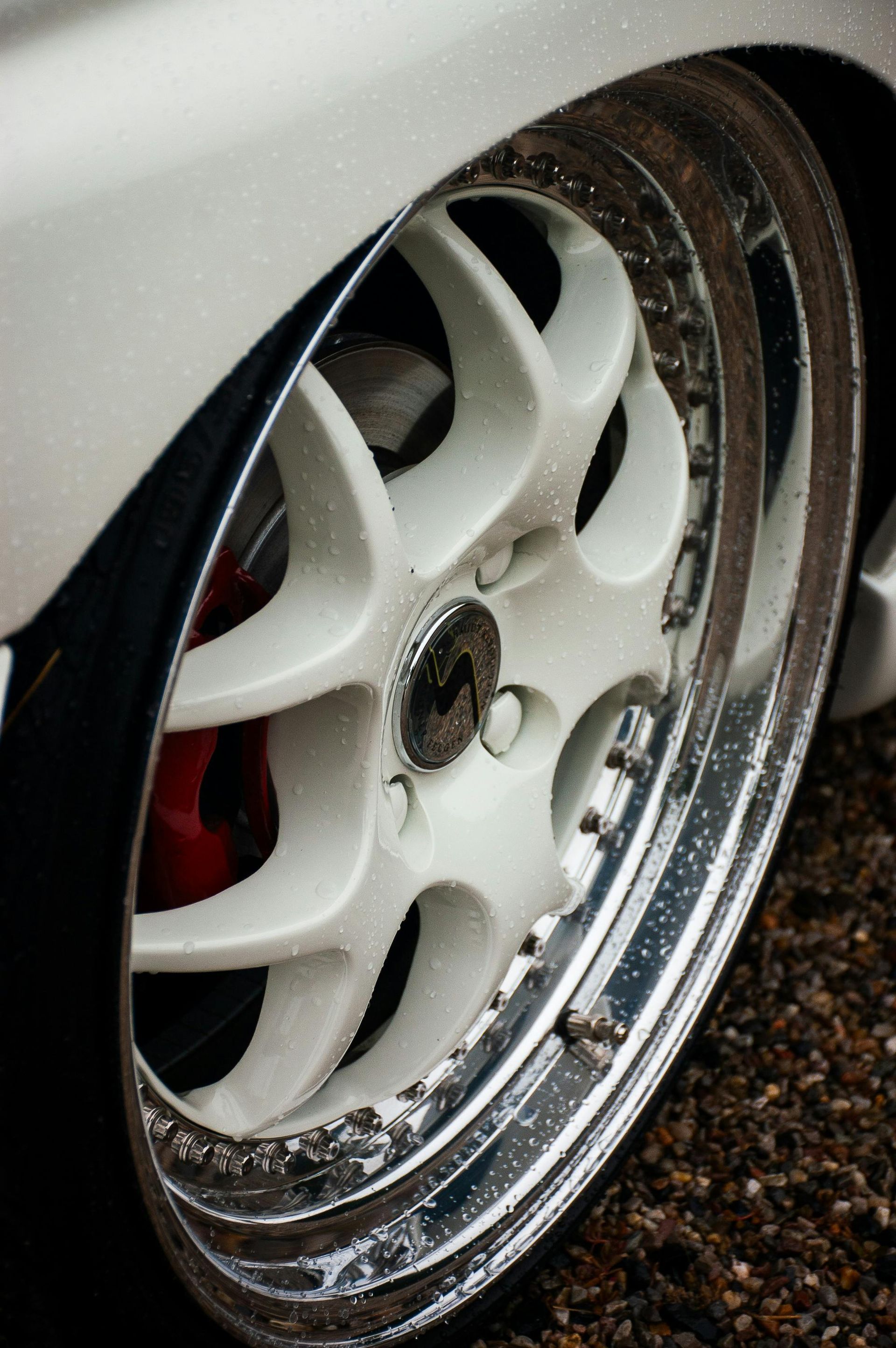 White car wheel with a chrome rim, against a blurred background. Red brake caliper visible.
