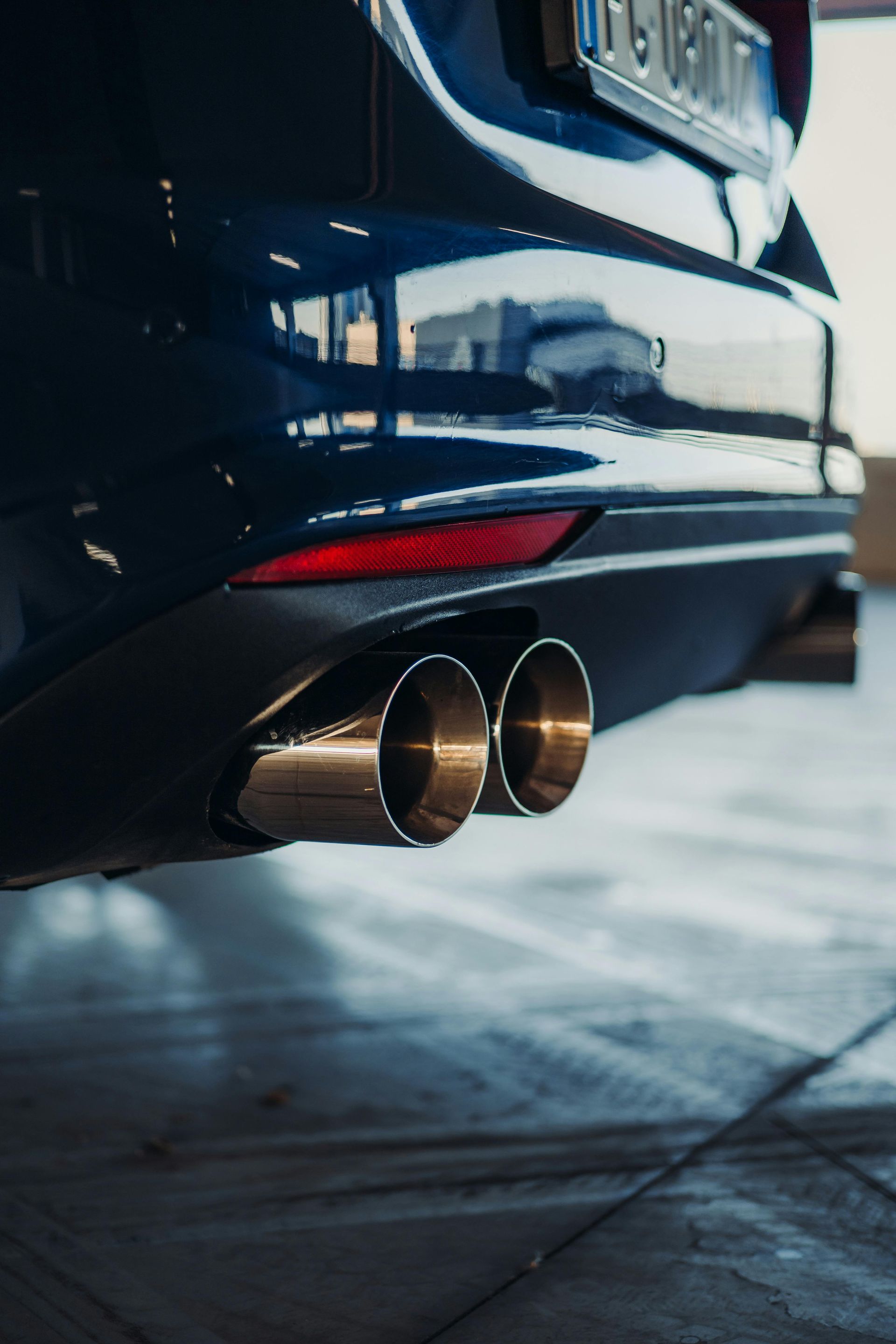 Close-up of a car's rear end with shiny exhaust pipes. Blue paint, red reflector, and a dark, textured setting.